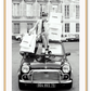 A black and white vintage fashion wall art with a lady holding Chanel shopping bags riding on vintage car Fiat in European city. 