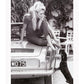 A black and white photo print of a fashion model woman lady on vintage car patting dog dachshund blonde and sexy 